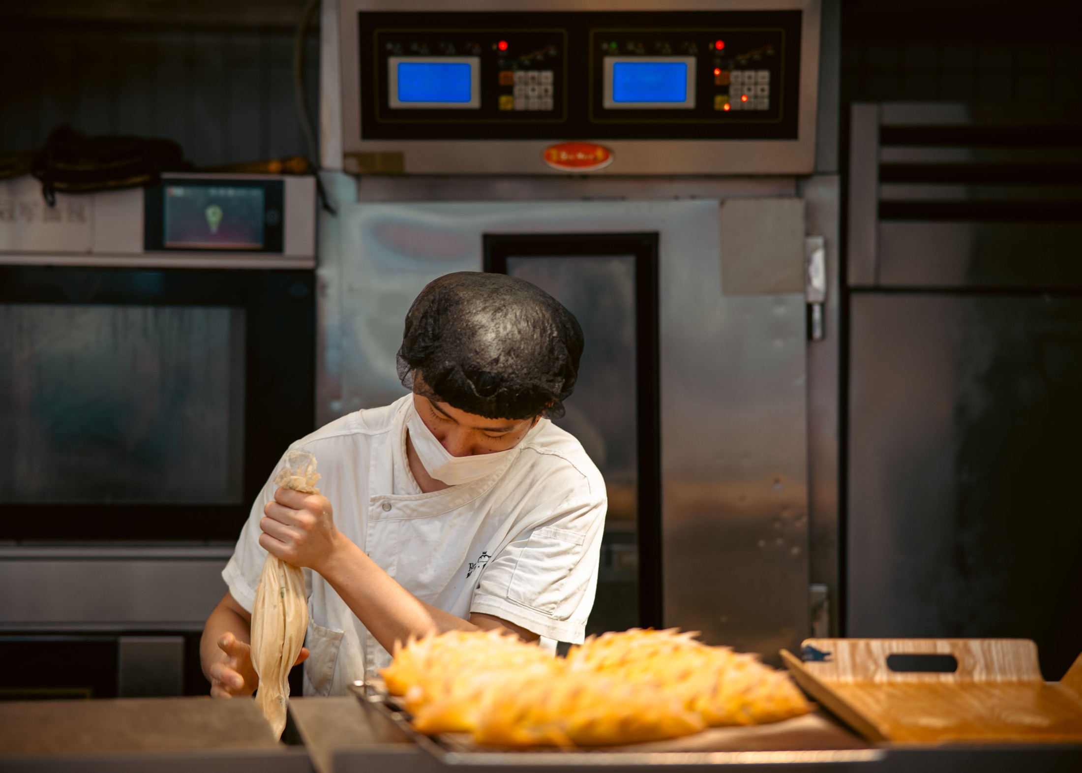 A masked pastry chef using a piping bag at a counter in front of bakery ovens.