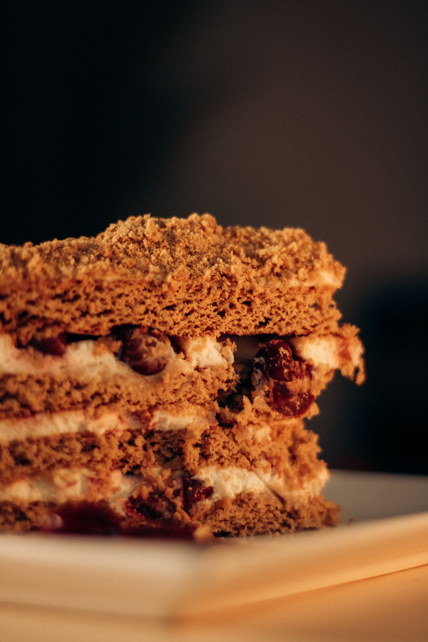 A cut layered cake with cream and dark fruit on a plate against a dark background.