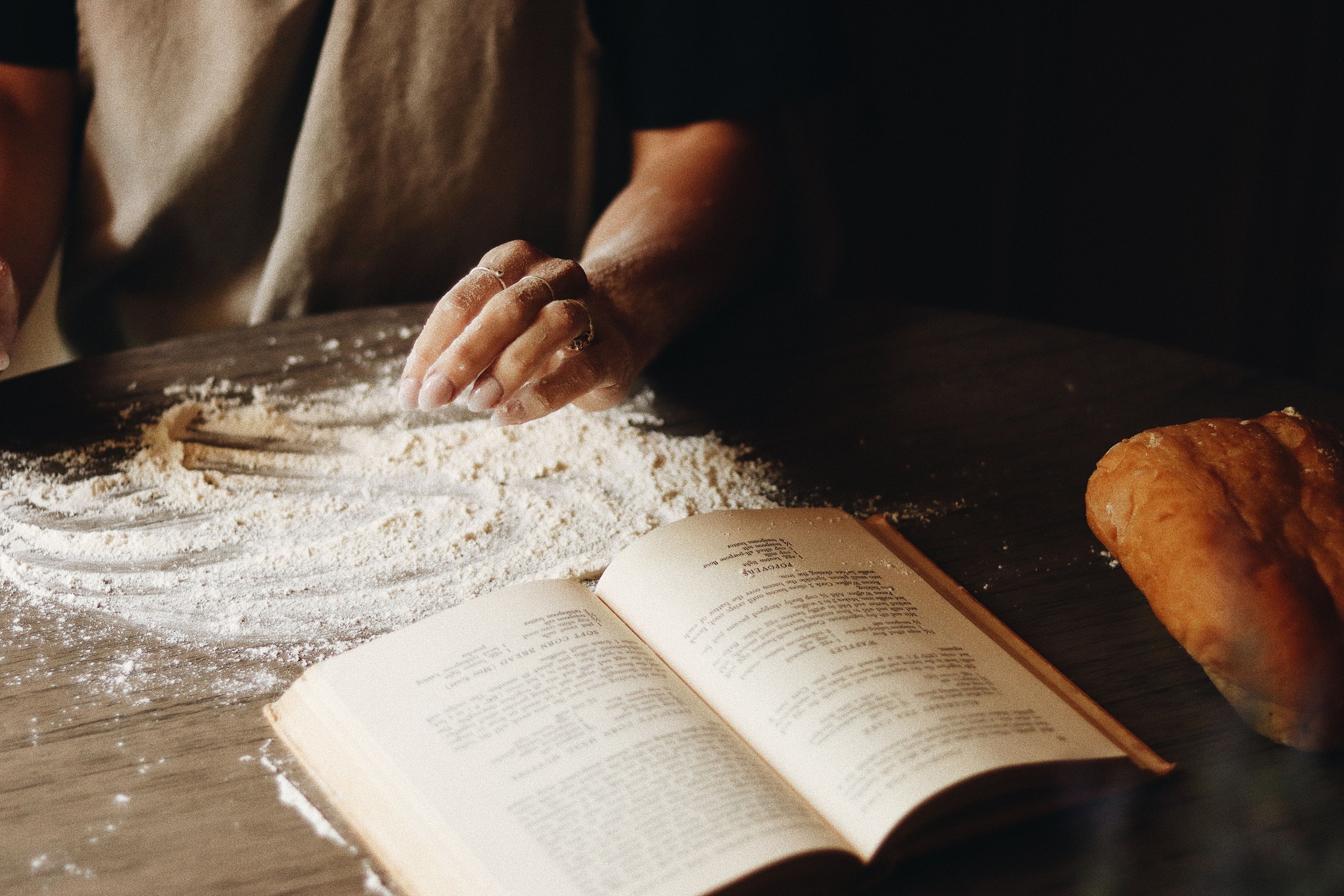 An open recipe book beside flour, dough and a loaf on a wooden work surface.