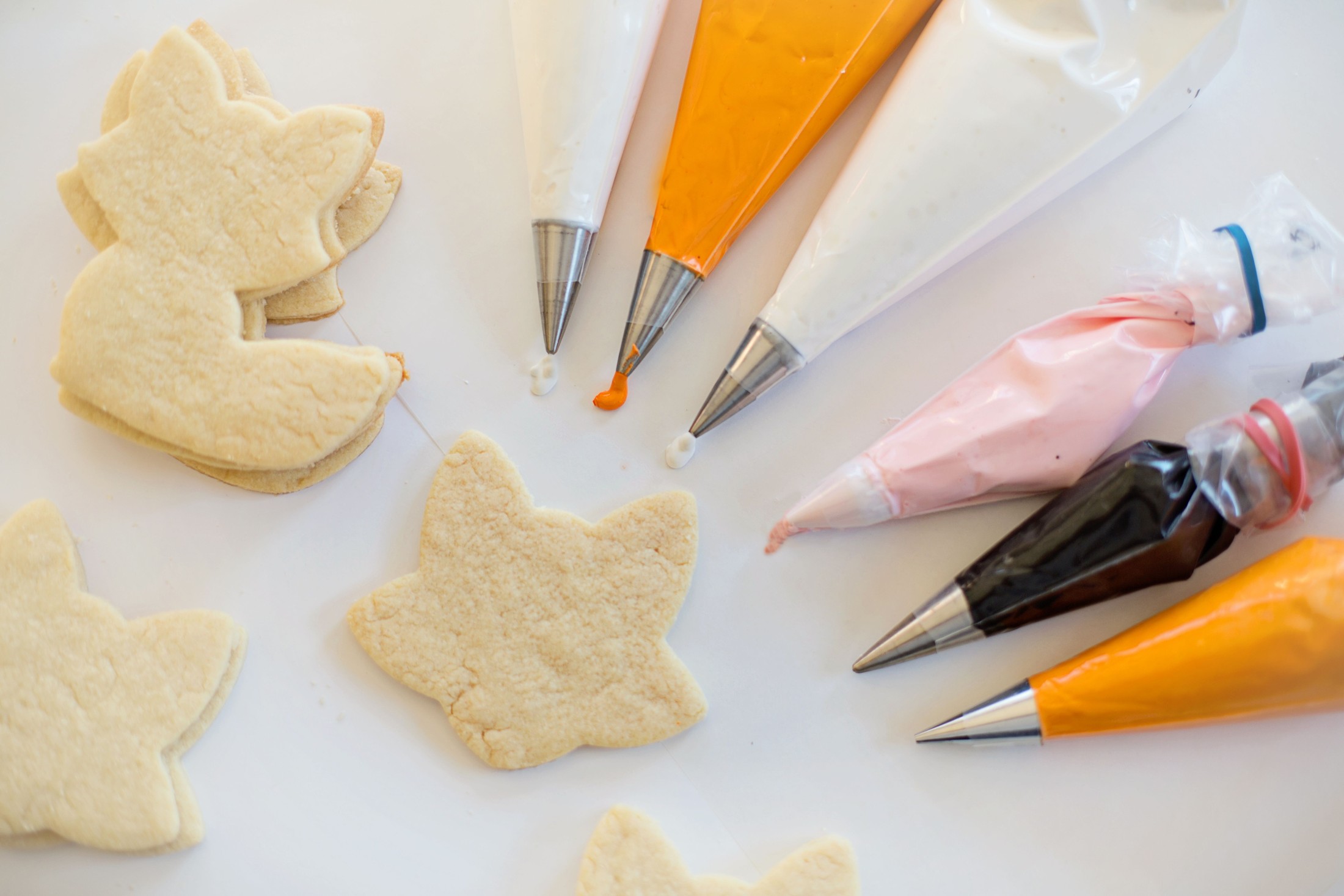 Coloured icing bags with metal tips beside leaf-shaped cookies on a white surface.